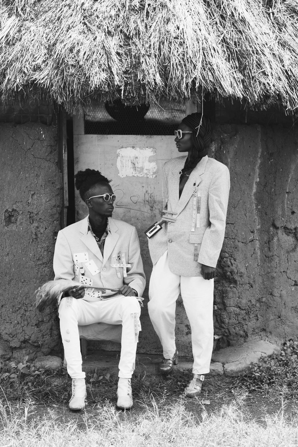 Black and white contemporary African art portrait of two men in modern tailored suits beneath a thatched hut, symbolising heritage and style.