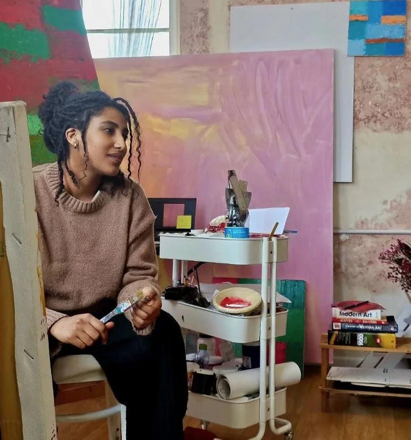 Helen Zeray, a contemporary African artist, sitting in her studio holding a paintbrush. The space features large colorful canvases and art supplies